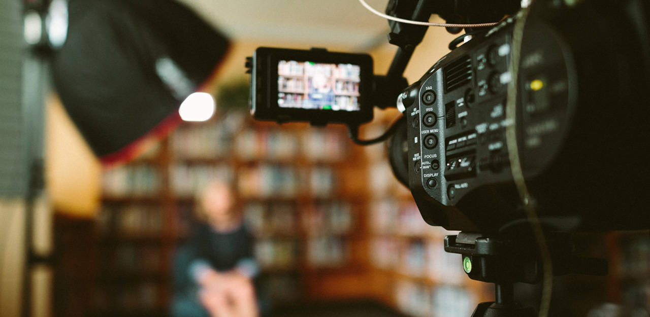 A person sitting in front of a bookshelf, being recorded on video.