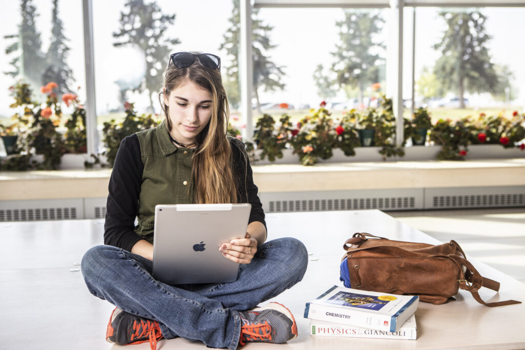 A student sitting cross-legged in a sunny room, typing on her laptop.
