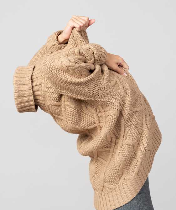 Woman pulling a cable-knit beige sweater over her head.