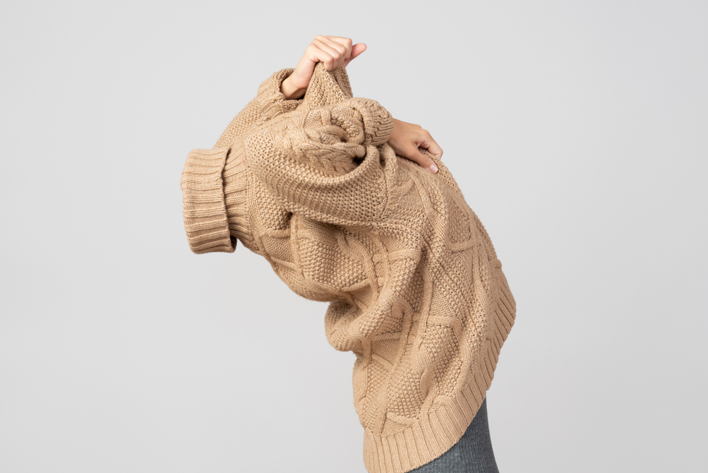Woman pulling a cable-knit beige sweater over her head.
