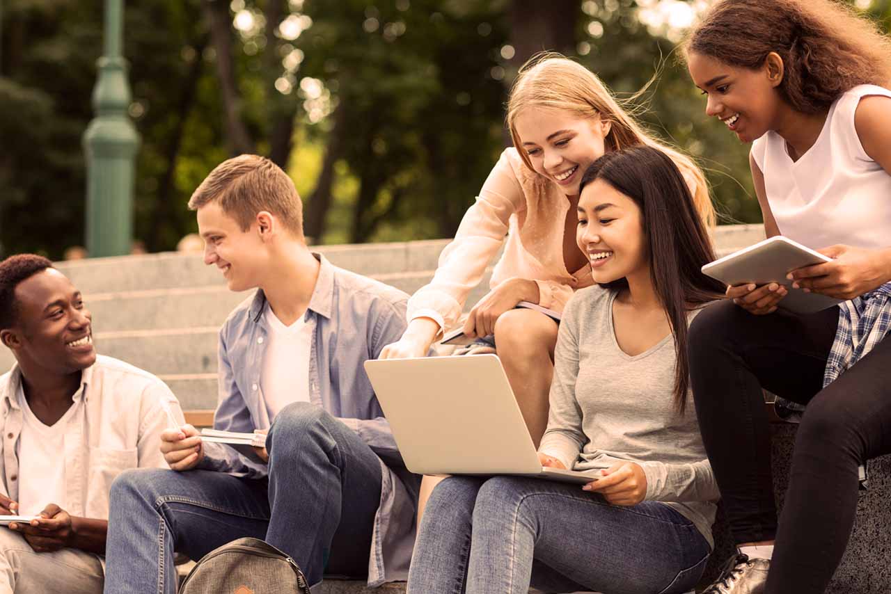 Young adult students sitting outside and working on a project. One student holds a laptop.