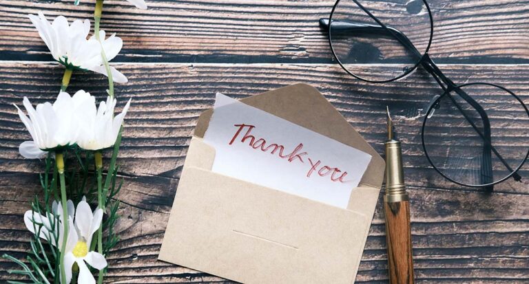 A flat-lay arrangement featuring a thank you card, a fountain pen, glasses, and flowers, all on a distressed wood desktop.